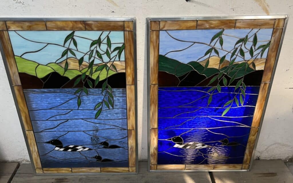 A pair of rectangular stained glass panels depicting a serene lake scene. The left panel shows a lighter blue lake with an adult loon and two small loons, while the right panel features a darker blue lake with an adult loon and one small loon. Both panels include light blue skies, green and tan hills, and green hanging willow-like branches. The panels are framed in metallic came.