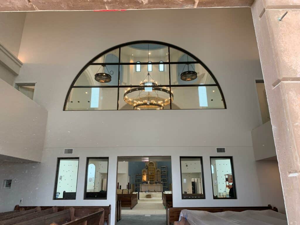 A large, semi-circular clearstory window with black grid lines, set high in a light-colored wall, reflecting three ornate chandeliers in a church or sanctuary interior.