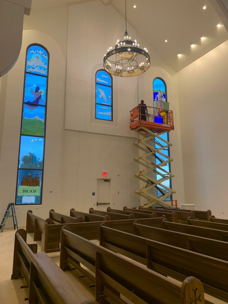 An interior view of a bright church sanctuary where a worker on an orange scissor lift is installing or maintaining an arched stained glass window. Another tall stained glass window on the left depicts multiple religious scenes including figures, landscapes, and a building marked '301 A.D.'. Wooden pews are arranged in the foreground, and a large, ornate metal chandelier hangs from the high white ceiling.