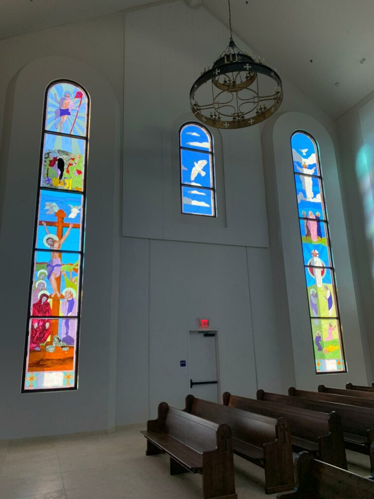 A church interior with three tall, arched stained glass windows depicting Christian narratives including the Crucifixion, Resurrection, Baptism, and the Holy Spirit dove, flanked by rows of wooden pews.