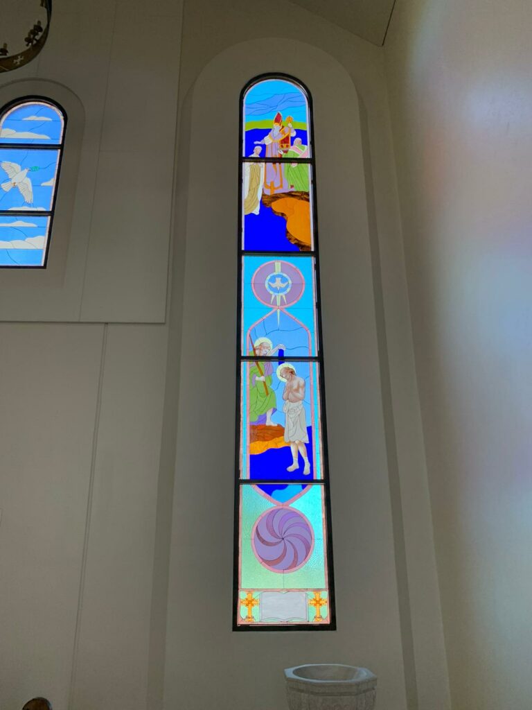 A tall, arched multi-paneled stained glass window depicting biblical scenes, including a dove symbol and the baptism of Christ, with a swirling geometric design at the bottom.