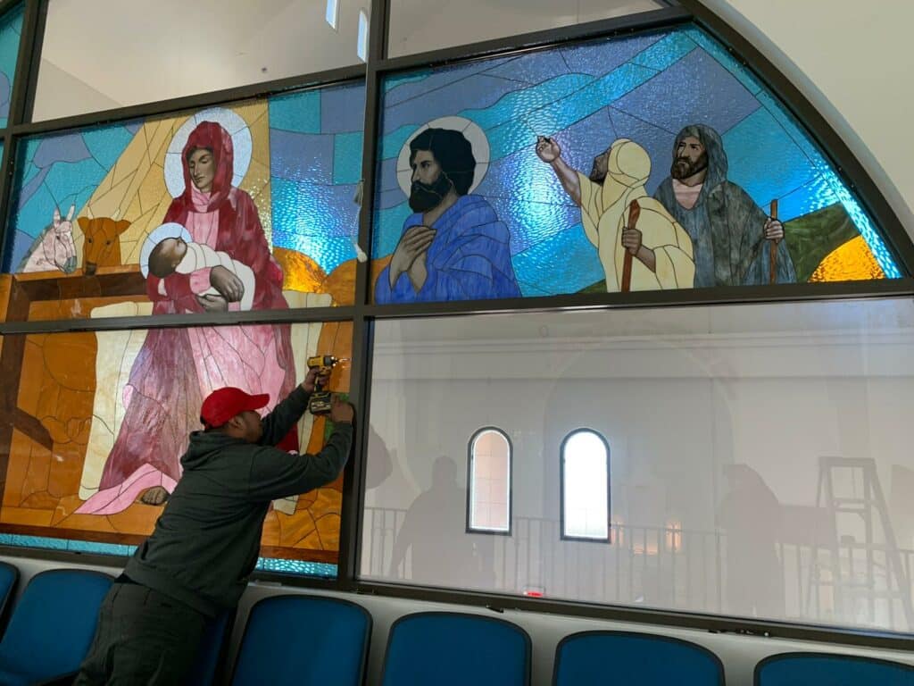 A craftsman uses a drill to install large, framed stained glass panels in a church setting. The panels depict biblical figures: the Virgin Mary holding baby Jesus, a robed man with a halo, and two prophets, one pointing heavenward. Below the vibrant religious panels are clear windows showing the church interior with arched openings.