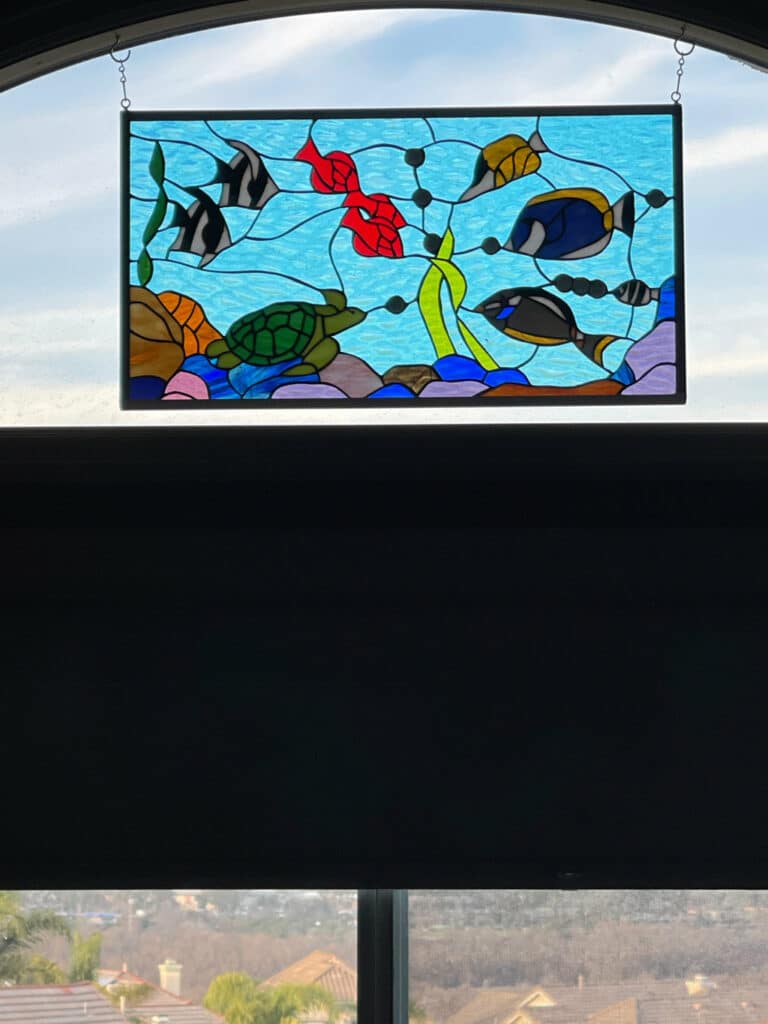 A rectangular stained glass panel depicting an underwater scene with a green sea turtle, black and white angelfish, red coral, blue tang fish, and green seaweed against a light blue water background, suspended in an arched window opening.