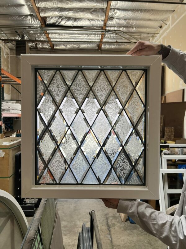 A square, insulated stained glass panel with a classic diamond pattern. The panel features alternating clear beveled diamonds and clear textured glass diamonds, creating a sparkling and private effect. It is framed with black lead lines and set within a clean white vinyl frame, held by a person's hands in a workshop.