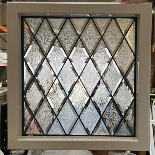 A square, insulated stained glass panel with a classic diamond pattern. The panel features alternating clear beveled diamonds and clear textured glass diamonds, creating a sparkling and private effect. It is framed with black lead lines and set within a clean white vinyl frame, held by a person's hands in a workshop.