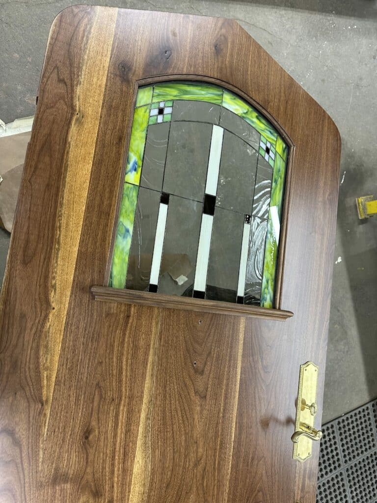 A newly crafted solid walnut entry door, featuring an arched leaded stained glass panel. The glass design includes vibrant green swirled glass, clear textured glass, opaque white rectangular strips, and small black squares arranged in a geometric, Craftsman style. A classic brass door handle set is visible on the richly grained wooden door.