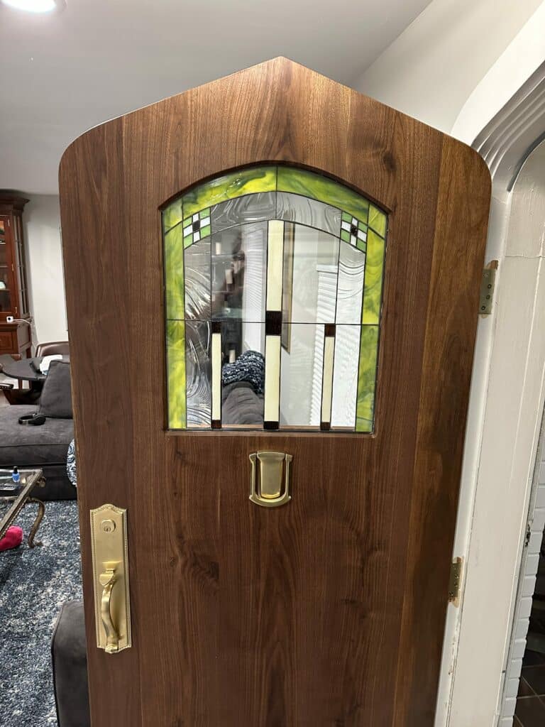 A rich, dark wood arched entry door featuring an integrated leaded stained glass panel with a geometric Prairie-style design. The stained glass incorporates vibrant chartreuse green, clear textured glass, and cream accents, set within dark leading. Brass hardware including a mail slot and handle is visible on the door.