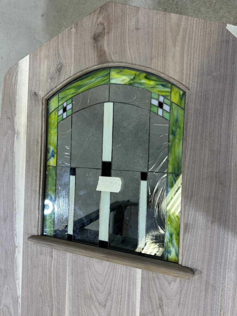 An arched leaded stained glass panel with a geometric design, set into a natural, light brown wooden door. The stained glass features a mottled green and yellow border, clear textured glass sections, and a central pattern of vertical white opalescent glass bars and small black and white square accents. A small wooden sill is present beneath the glass panel.