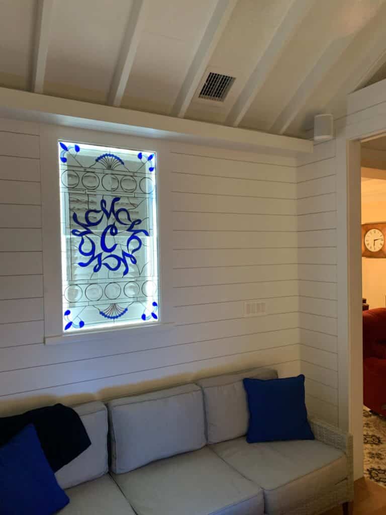 A rectangular leaded stained glass panel featuring an intricate, vibrant blue scrolling design at its center, surrounded by clear textured glass with circular elements and blue fan-like motifs at the top and bottom. The panel is backlit and installed in a white shiplap wall above a gray sofa.