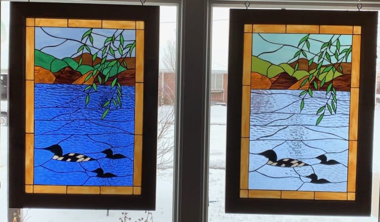 A pair of complementary rectangular stained glass panels depicting a serene lake. Each panel features a black and white loon with two ducklings swimming in textured blue water, bordered by green and brown hills under a light blue sky, with green leafy branches draping from above. The left panel uses deeper blues and greens, while the right features lighter, softer hues.