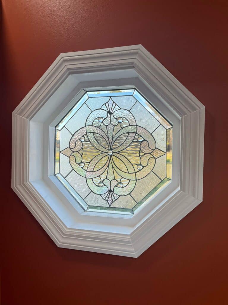 An octagonal leaded stained glass window panel installed in a deep red wall. The clear glass design features a central ornate floral medallion surrounded by scrollwork, crafted with a mix of textured glass, small round bevels, and larger straight beveled pieces. Sunlight filters through, showing trees outside.