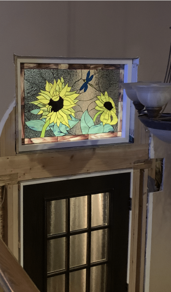 Rectangular stained glass transom panel featuring two vibrant yellow sunflowers, green leaves, and a blue dragonfly on a textured clear and beige background, installed above a black entry door.