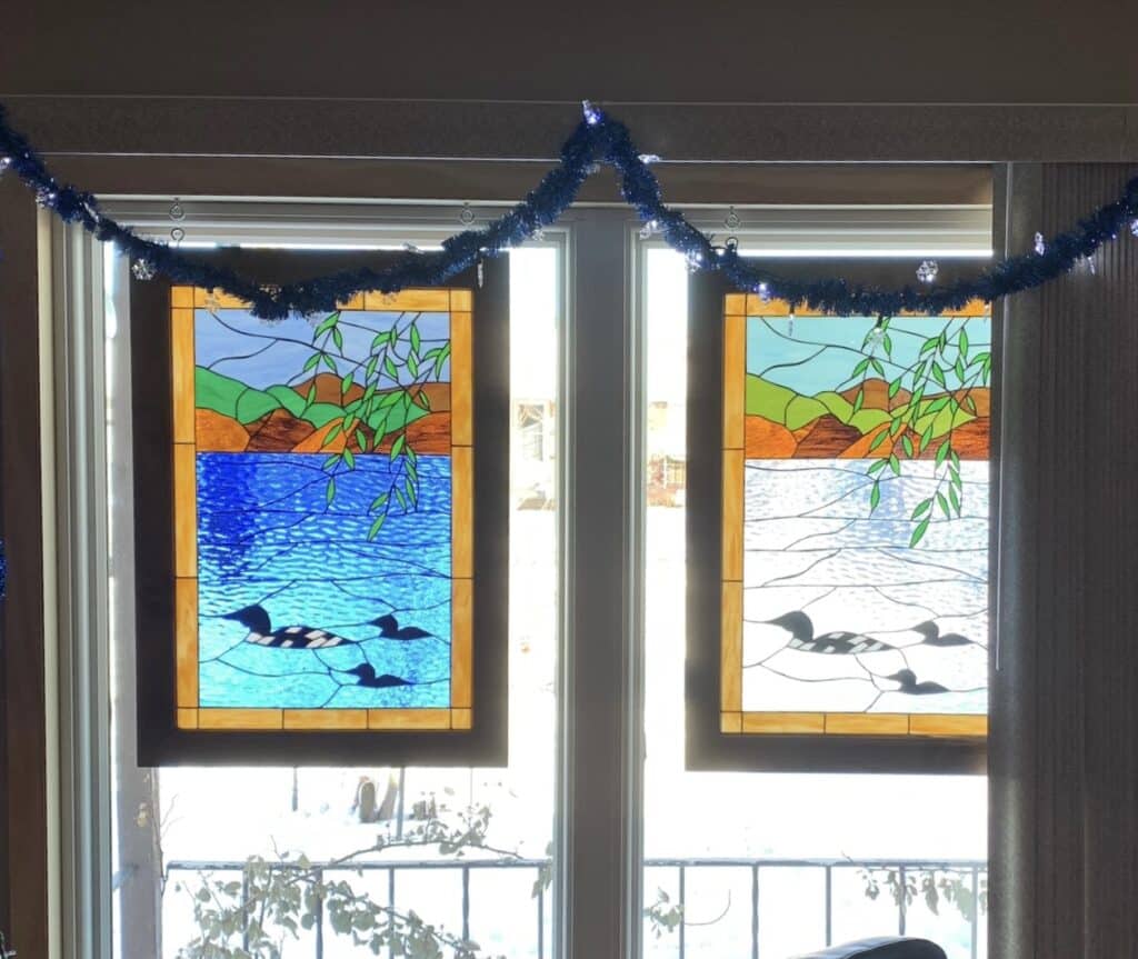 Two rectangular stained glass panels hung in a window, depicting a serene lakeside scene. Each panel features a large adult loon swimming with two smaller loon chicks in textured water. The left panel shows vibrant blue water, while the right panel features lighter, icy-blue water. Both panels include a background of rolling green and brown hills under a light blue sky, with green willow-like branches descending from the top. Dark frames surround each panel.