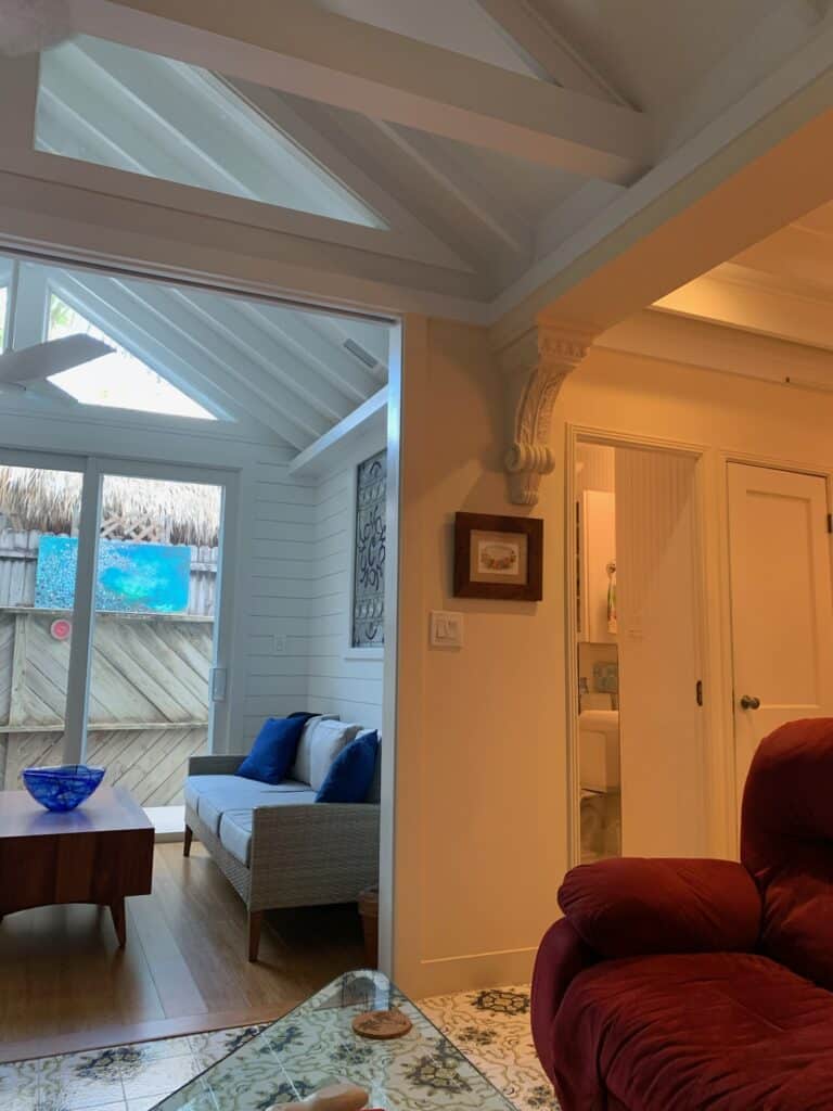 An interior living room with a decorative rectangular leaded glass panel inset into a white shiplap wall, featuring an ornate scroll design. The room includes a sliding glass door, a wicker sofa with blue pillows, and high triangular transom windows. A red armchair and patterned floor tiles are visible in the foreground.