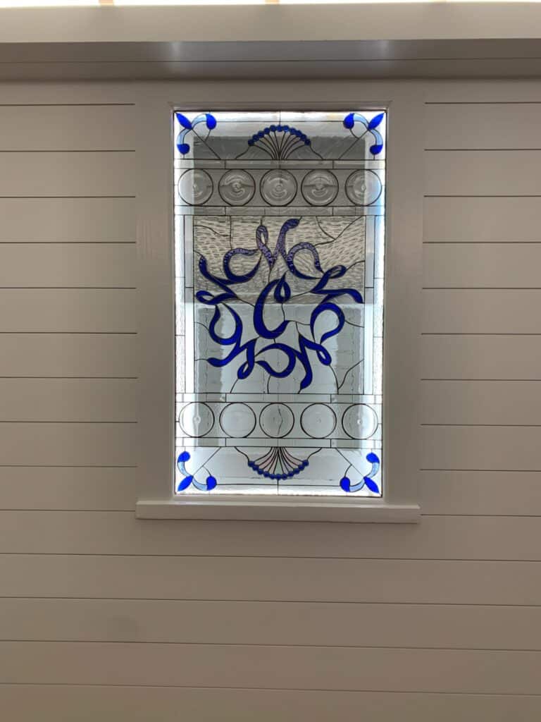 A rectangular leaded stained glass window panel featuring an ornate, swirling blue monogram resembling the letter 'M' at its center. The monogram is surrounded by clear textured glass, circular bevels, and smaller blue scroll patterns in the corners and along the top and bottom edges. The panel is installed in a light gray shiplap wall.