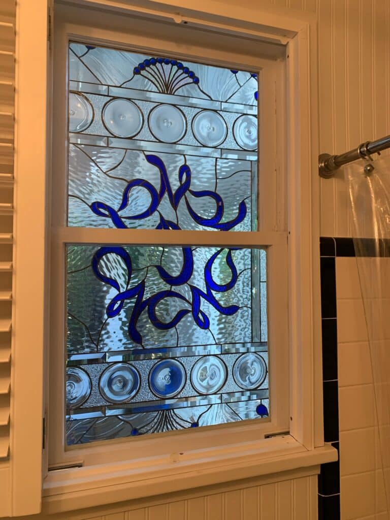 A rectangular stained glass window in a white frame, featuring a dynamic design of flowing, abstract blue glass swirls set against a backdrop of clear, highly textured and beveled glass. The top and bottom sections include horizontal bands with clear glass rondels/jewels and a delicate blue fan accent at the top center. The window is installed in a bathroom setting with white beadboard paneling and white tiled walls with a black trim.