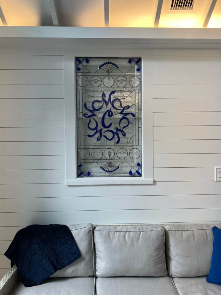 A rectangular stained glass panel featuring a prominent, abstract swirling pattern in royal blue glass. The design is complemented by clear, textured, and bevel-cut glass segments, including circular rondels, and smaller blue fan-like elements at the top and bottom. The panel is framed in white and installed in a white shiplap wall above a light grey sofa.