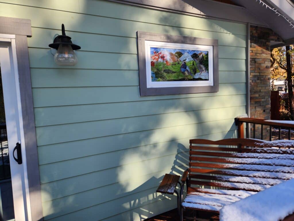 A rectangular stained glass panel mounted on light green horizontal siding, featuring a vibrant nature scene with two blue and gray birds, red poppy-like flowers, green rolling hills, a winding path, and blue sky. The panel is set within a gray exterior frame and a white inner frame.