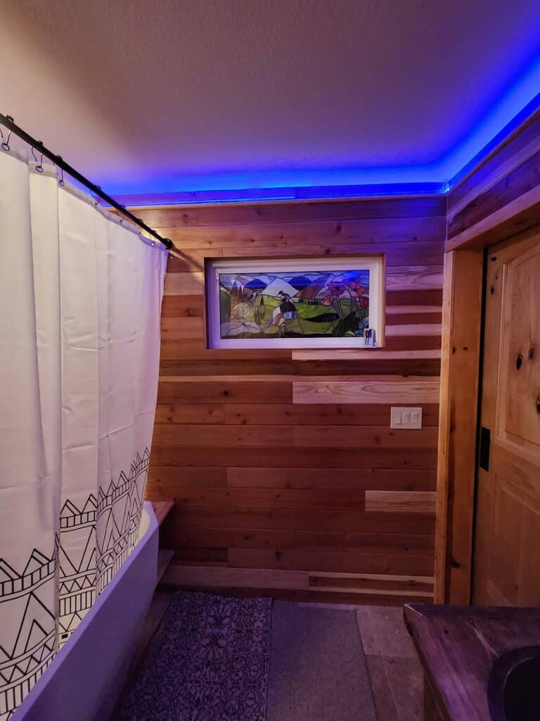 A rectangular leaded stained glass panel depicting two magpie-like birds, red poppies, and purple mountains, set into a warm cedar-paneled wall in a rustic bathroom. The room features blue LED strip lighting along the ceiling and a white shower curtain with a black geometric pattern.