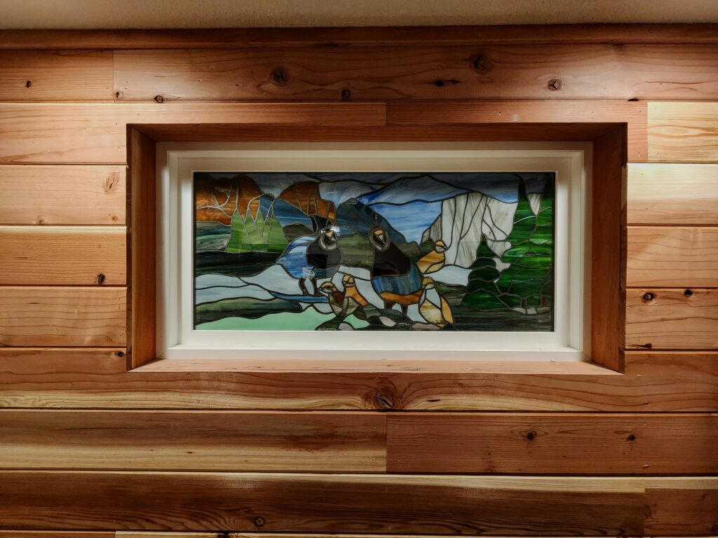 A rectangular stained glass panel depicting two dark-bodied adult seals and several lighter-colored seal pups resting on blue-grey ice or rocks. The background features blue and grey jagged mountains, brown hills, and areas of dark green and blue water. The panel is set in a white frame, which is recessed into a wall made of light-colored, horizontal wooden planks.