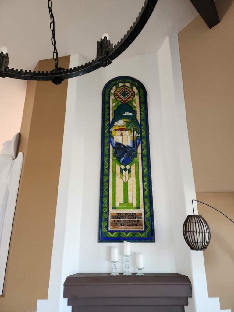 A tall, arched stained glass panel depicting a vibrant, Southwestern-style landscape with a mission building, palm trees, and blue water, topped by a geometric design featuring an eye. Below the landscape, the panel displays an inscription in dark letters: 'TO YOUR DESCENDANTS I WILL GIVE THIS LAND'. The panel is framed by intricate green, blue, and amber geometric patterns and is mounted on a white wall above a dark brown mantelpiece with three white candles.