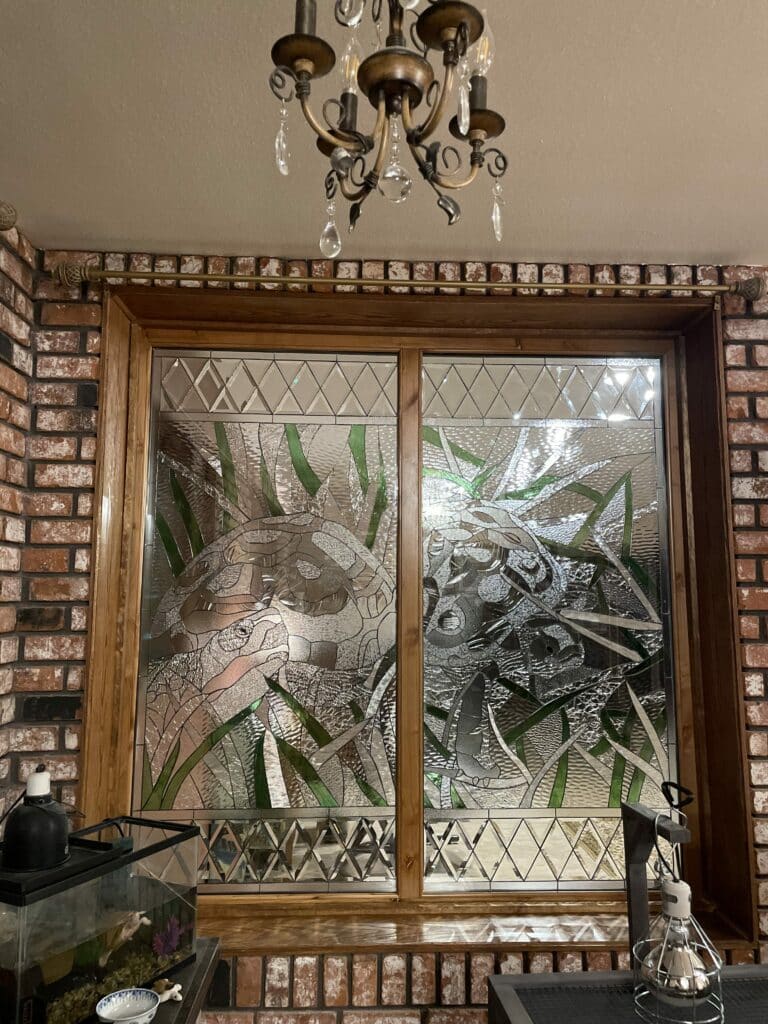 A large rectangular leaded stained glass window, set in a wooden frame. The central design depicts two detailed turtles swimming among tall green reeds, using various clear textured glass for the turtles and water, and translucent green glass for the plants. The top and bottom sections feature decorative borders of clear beveled diamond and square shaped glass.