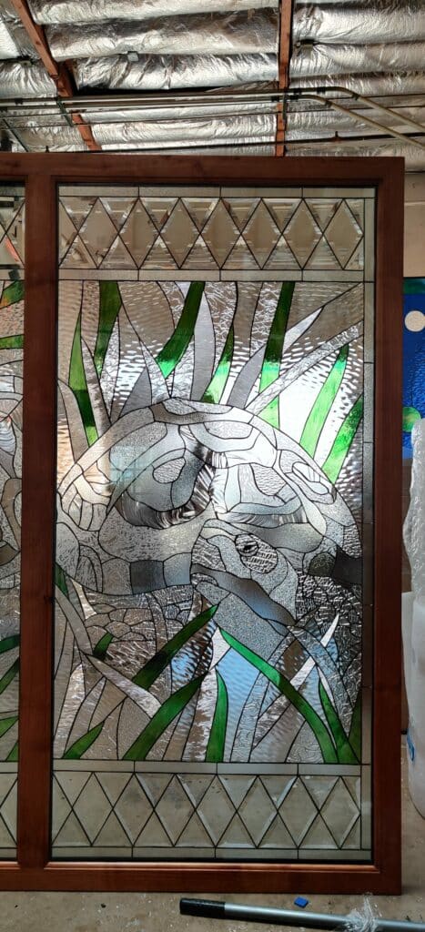 A rectangular leaded stained glass panel framed in dark wood, featuring a larger and smaller turtle swimming amidst tall green reeds and clear textured water. The main scene is bordered top and bottom with clear beveled diamonds set against textured clear glass.