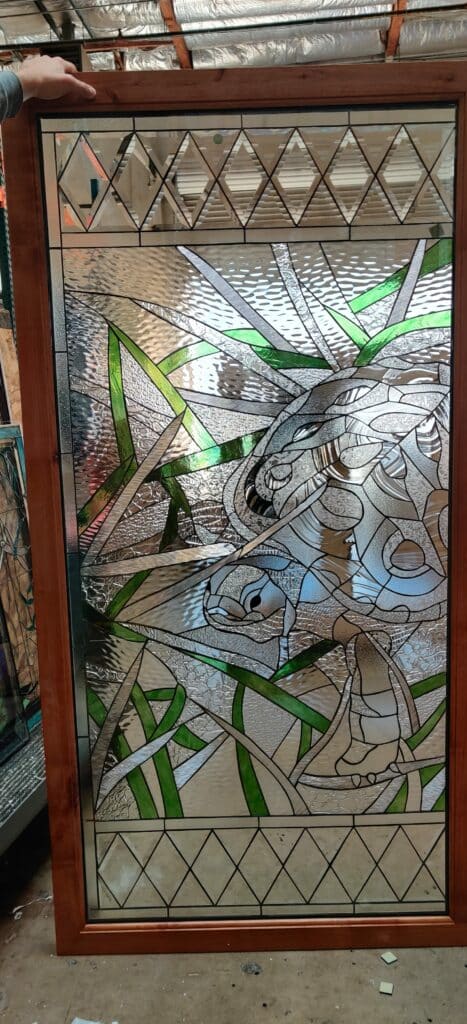 A rectangular stained glass panel within a warm brown wooden frame, depicting a sea turtle swimming amidst vibrant green reeds and textured clear water. The turtle's shell, head, and flippers are formed from various clear, white, and subtly rippled textured glass pieces. The upper and lower borders of the panel feature clear beveled diamond-shaped glass segments.