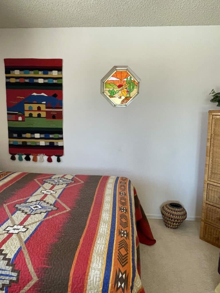 An octagonal stained glass panel in a metallic frame, depicting a desert scene at sunset. The vibrant sky features orange and yellow hues radiating from a setting sun, illuminating green saguaro cacti and other desert plants. A stylized figure in yellow clothing with a sombrero is seated on the brown and green landscape.