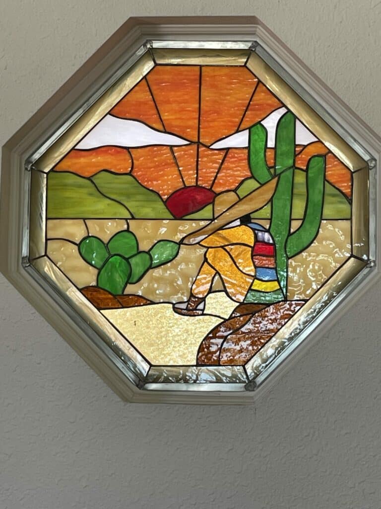 An octagonal stained glass window depicting a desert scene at sunset, featuring a person wearing a sombrero resting against a saguaro cactus, with a prickly pear cactus, distant mountains, and a vibrant orange and red sky.