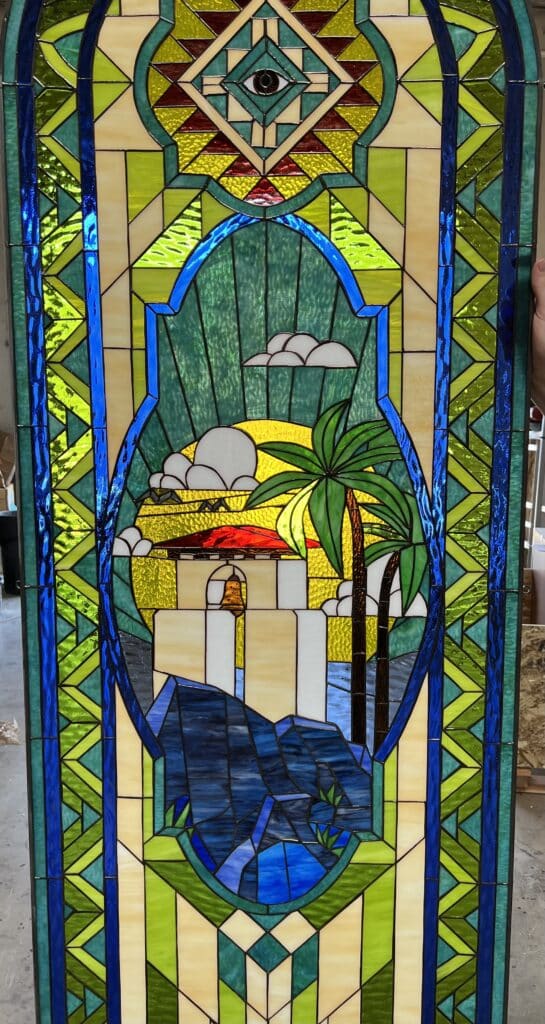 A rectangular leaded stained glass panel depicting a vibrant coastal mission scene within an oval vignette. The central image features a white building with a red roof and bell tower on a blue rocky coastline, with two green palm trees, a bright yellow sun, and white clouds. The scene is bordered by intricate geometric patterns in shades of emerald, lime, and deep blue, with cream accents. At the very top, a prominent Eye of Providence is set within a diamond motif.