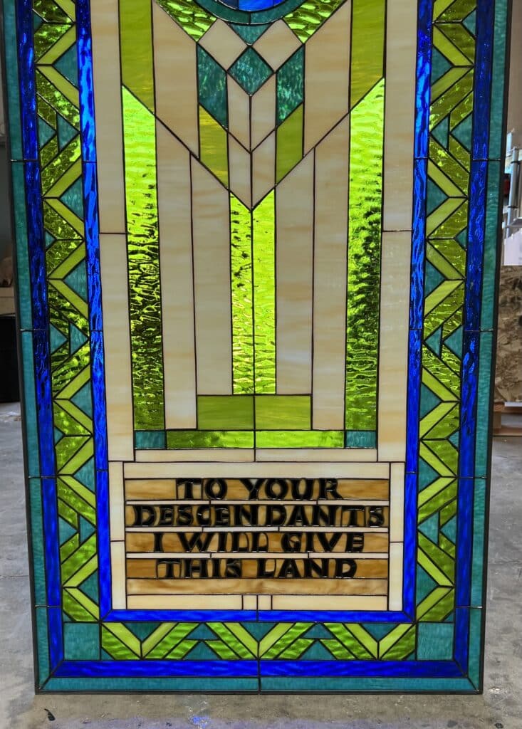 A rectangular leaded stained glass panel with a vibrant geometric design in shades of lime green, teal, and cobalt blue. A large, stylized 'M' is central, formed by cream and bright green textured glass. Below it, a horizontal amber-cream panel displays the inscription 'TO YOUR DESCENDANTS I WILL GIVE THIS LAND' in black capital letters.