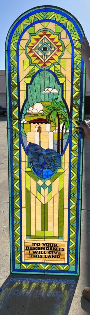 A tall, arched stained glass panel featuring a vibrant, multi-colored design. The top arch contains a geometric pattern in red, yellow, and green, centered around an 'Eye of Providence' symbol. Below, an oval medallion depicts a mission-style building with a bell, tall palm trees, fluffy clouds, and abstract blue water and mountains, all set against a yellow and green sky. The lower section includes a rectangular block with the text 'TO YOUR DESCENDANTS I WILL GIVE THIS LAND' in black lettering on amber glass. The entire panel is framed by intricate blue, green, and yellow geometric borders. Two hands are visible holding the panel against a sunny outdoor backdrop.