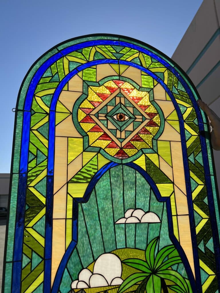 A vibrant, arched leaded stained glass panel. The design features a central 'Eye of Providence' in warm yellows and reds, surrounded by green and light cream geometric patterns and prominent blue arched bands. The lower portion depicts a verdant, textured green landscape with white clouds and a green palm frond. The panel is held up against a bright blue sky, showcasing its translucency and rich colors.