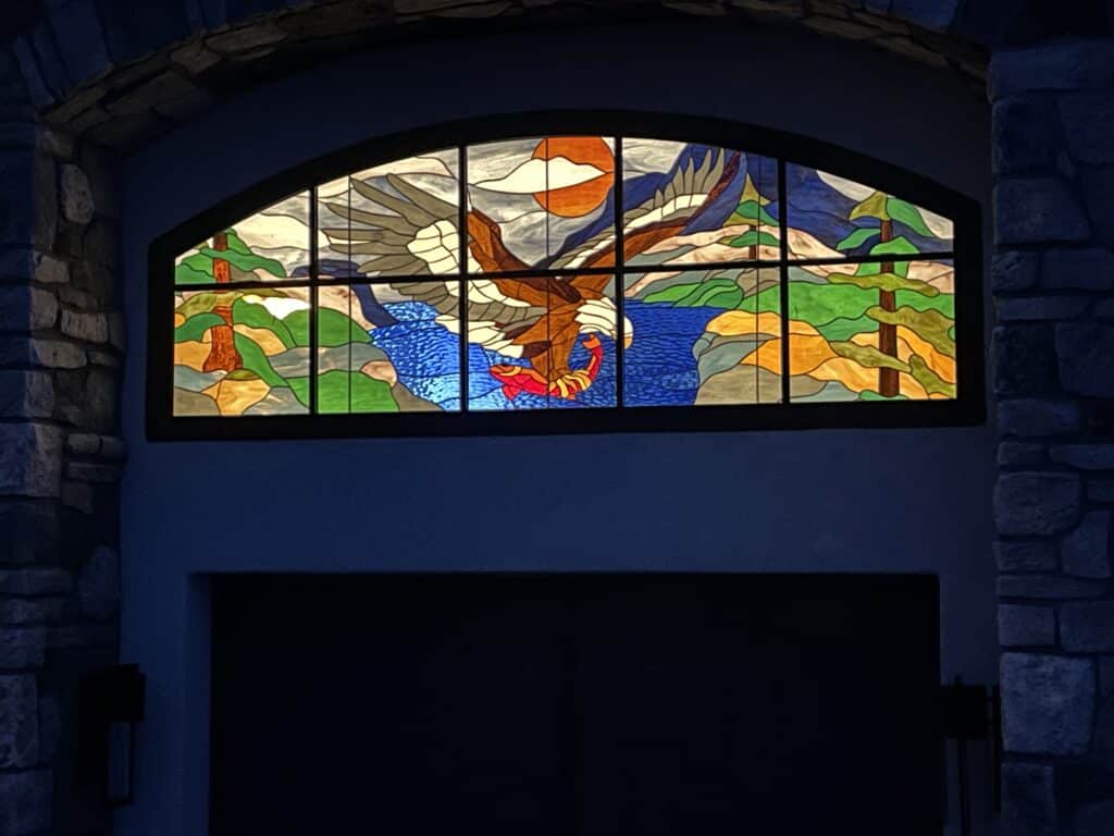 An arched leaded stained glass transom window depicting a majestic brown and white eagle with outstretched wings diving to catch a vibrant red salmon from a textured blue lake. The background includes a mountainous landscape with green pine trees and an orange setting or rising sun, all framed within a rustic stone archway.