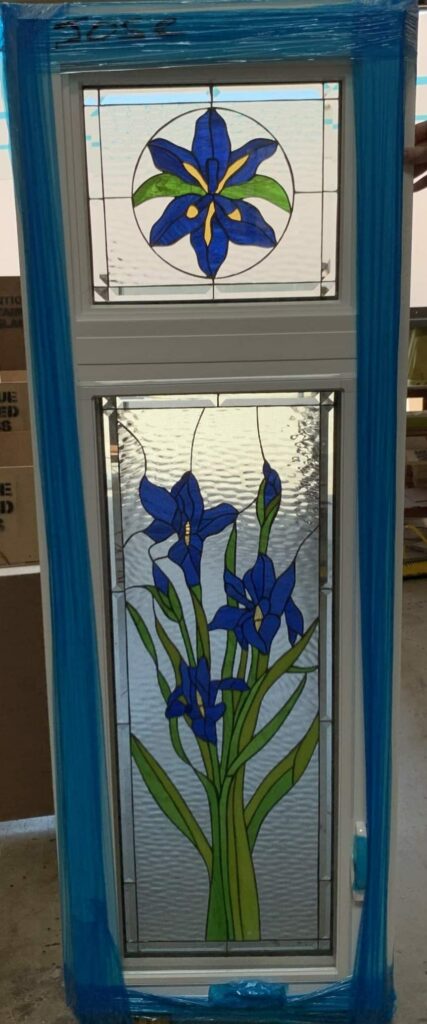 A tall, white-framed insulated window unit featuring two distinct stained glass panels. The upper panel displays a circular medallion with a single blue iris flower and green leaves. The larger lower panel showcases a vibrant vertical arrangement of multiple blue iris flowers, buds, and tall green stems, all set against clear textured and beveled glass. The window unit is wrapped in blue protective film and is shown in a warehouse setting.