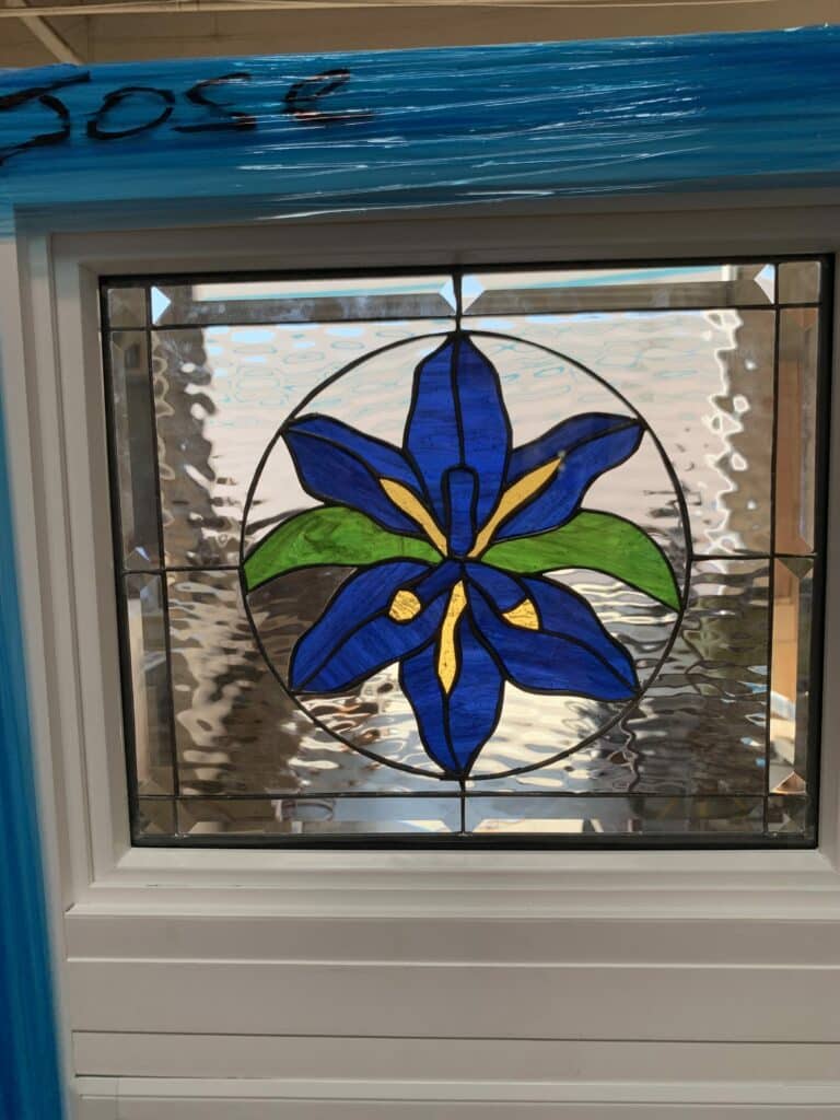 A rectangular stained glass window panel featuring a vibrant blue iris flower with green leaves and yellow accents, set within a circular clear textured glass background. The central medallion is surrounded by a border of clear rectangular and square beveled glass pieces, all framed in white.