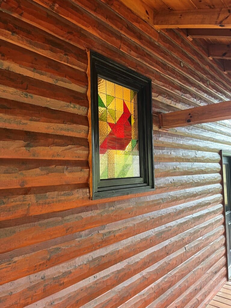 A rectangular stained glass window featuring a vibrant, geometric red cardinal with yellow and green abstract background shapes, set in a dark frame within a natural, rough-hewn log cabin wall.