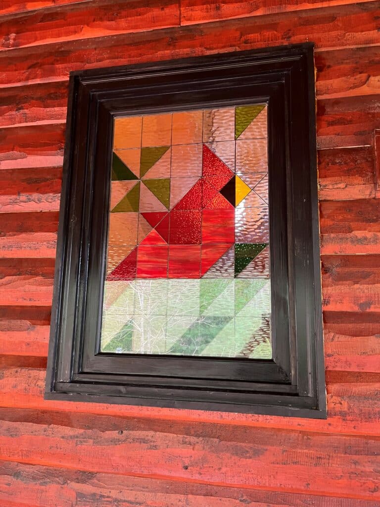 A rectangular stained glass window panel featuring a geometric, pixelated design of a red cardinal. The cardinal has a black eye and yellow beak, set against a background of clear, textured, and colored glass in shades of orange, green, and browns. The panel is encased in a dark wooden frame and mounted on a rustic reddish-orange wooden plank wall.