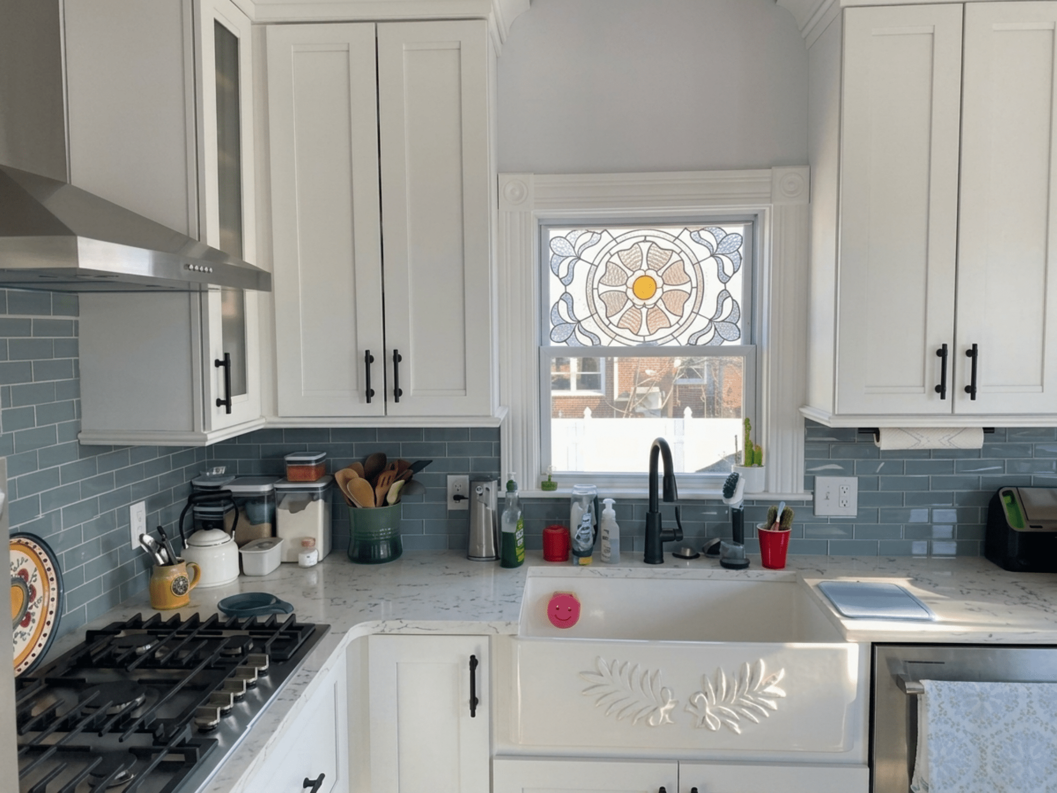 A rectangular stained glass panel depicting a central circular floral medallion. The medallion has a bright yellow core, encircled by an orange ring, then concentric layers of light brown and clear textured glass petals. Surrounding the medallion are symmetrical blue and clear textured geometric, leaf-like patterns, all against a clear textured background. The panel is installed in a white-framed kitchen window above a sink.