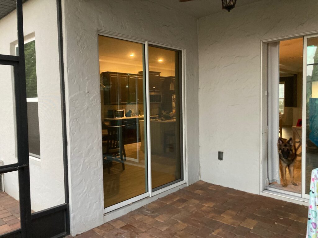 A large clear glass sliding door on a home patio reflects a kitchen with dark cabinets and warm lighting. Next to it, a smaller clear glass door shows a German Shepherd dog looking out. The patio has brick pavers, and the house walls are textured white stucco. A black screened enclosure is visible on the left.