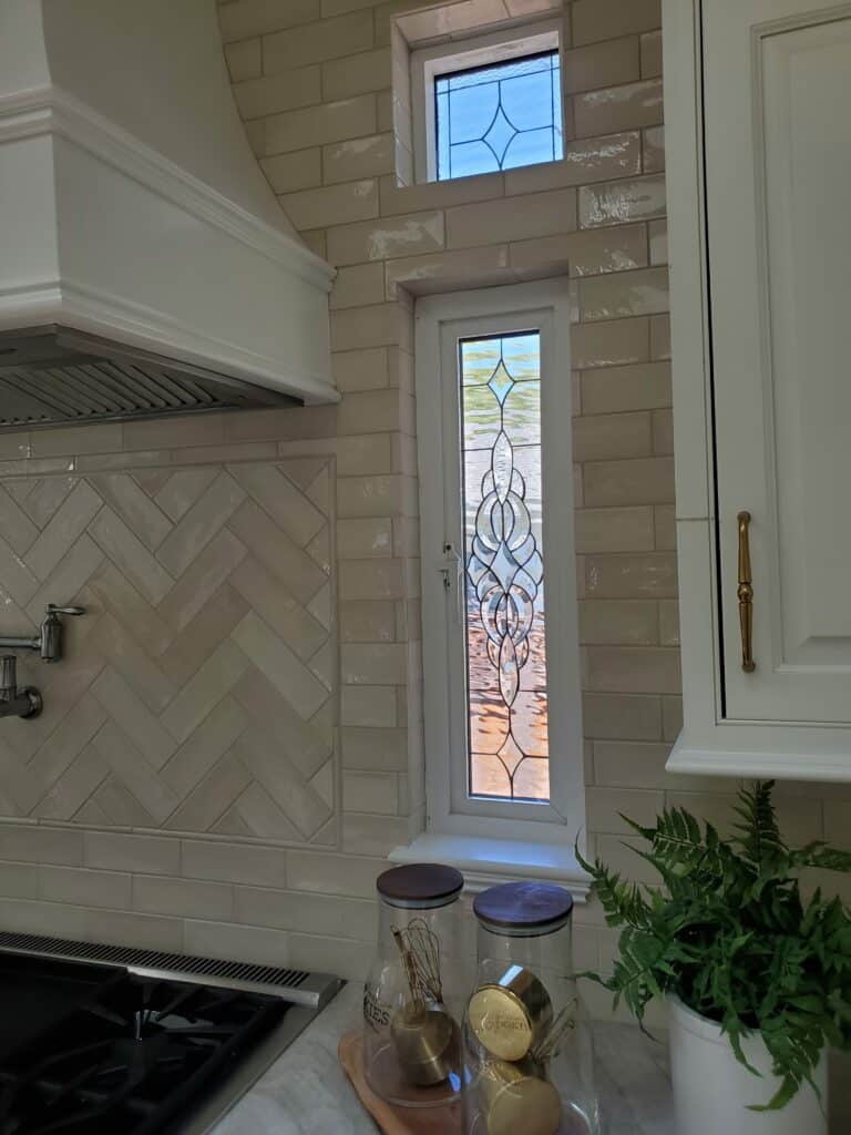 A modern kitchen features two custom clear beveled stained glass windows set into a beige subway tile wall. The lower, taller window has an intricate, flowing clear bevel design, while the smaller upper window has a simpler geometric clear bevel pattern. White cabinets, a range hood, and a gas range are also visible.