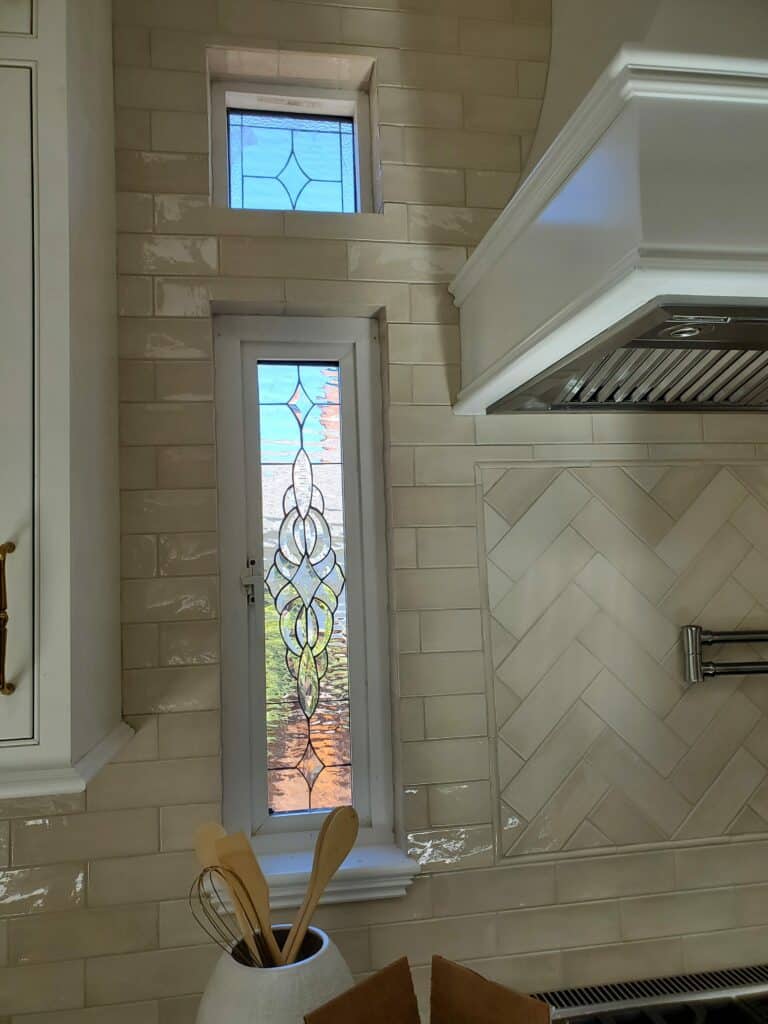 A modern kitchen interior featuring two white-framed rectangular clear beveled stained glass windows installed in a light cream subway tiled wall. The lower, taller window displays an intricate design of elegant scrolls and organic shapes, while the upper, smaller transom window features a simple geometric pattern with a central diamond.
