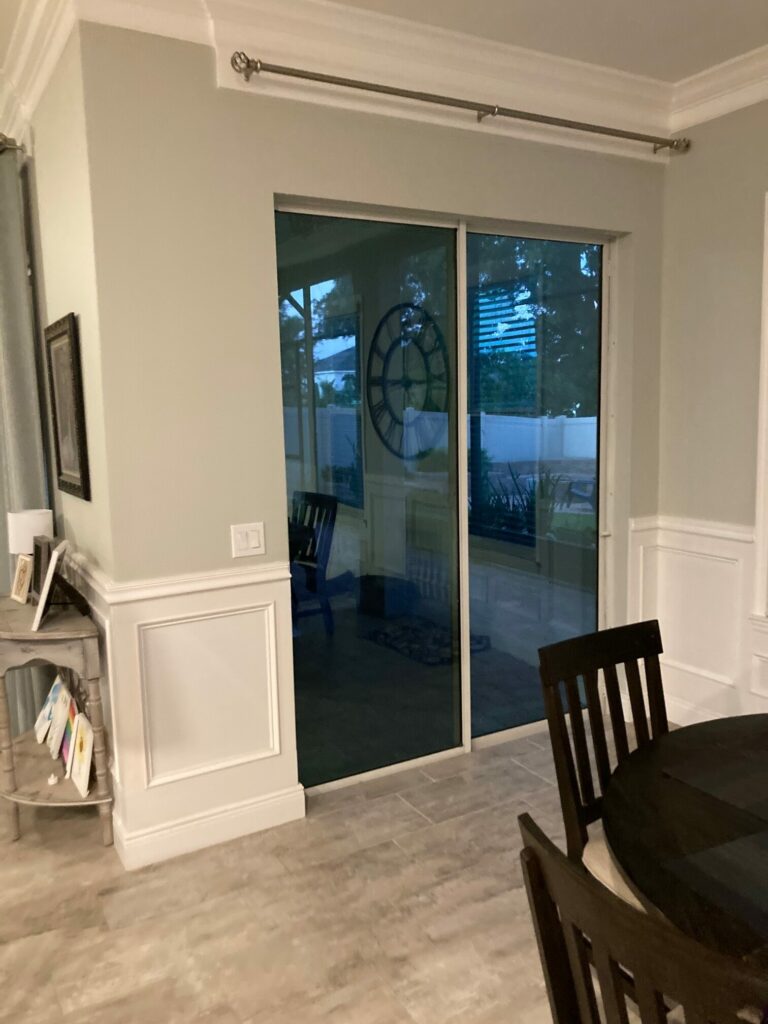 An interior shot of a dining room featuring light grey walls, white wainscoting, and large blue-tinted sliding glass doors looking out to a patio with a large wall clock visible, next to a dark wood dining table.