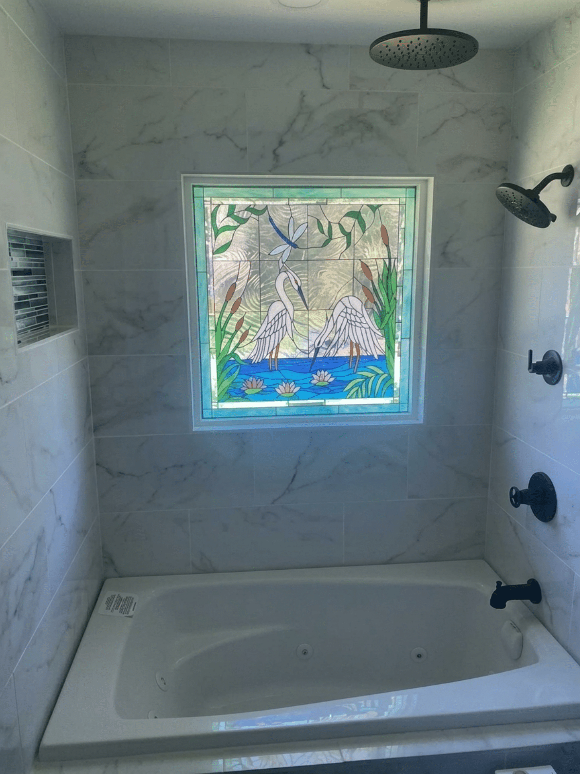 A rectangular stained glass panel installed in a white tiled bathroom, above a bathtub. The panel depicts two white herons wading in water with pink water lilies, green cattails, and a blue dragonfly. The background glass is textured and clear.