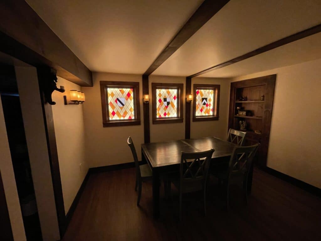 A cozy dining room showcasing three rectangular leaded stained glass windows, each with an amber, red, and clear diamond pattern and a central black, white, and red heraldic shield design, set in dark wood frames. The room features dark wood ceiling beams, matching dining table and chairs, wall sconces, and a built-in bookshelf, evoking a warm, traditional atmosphere.