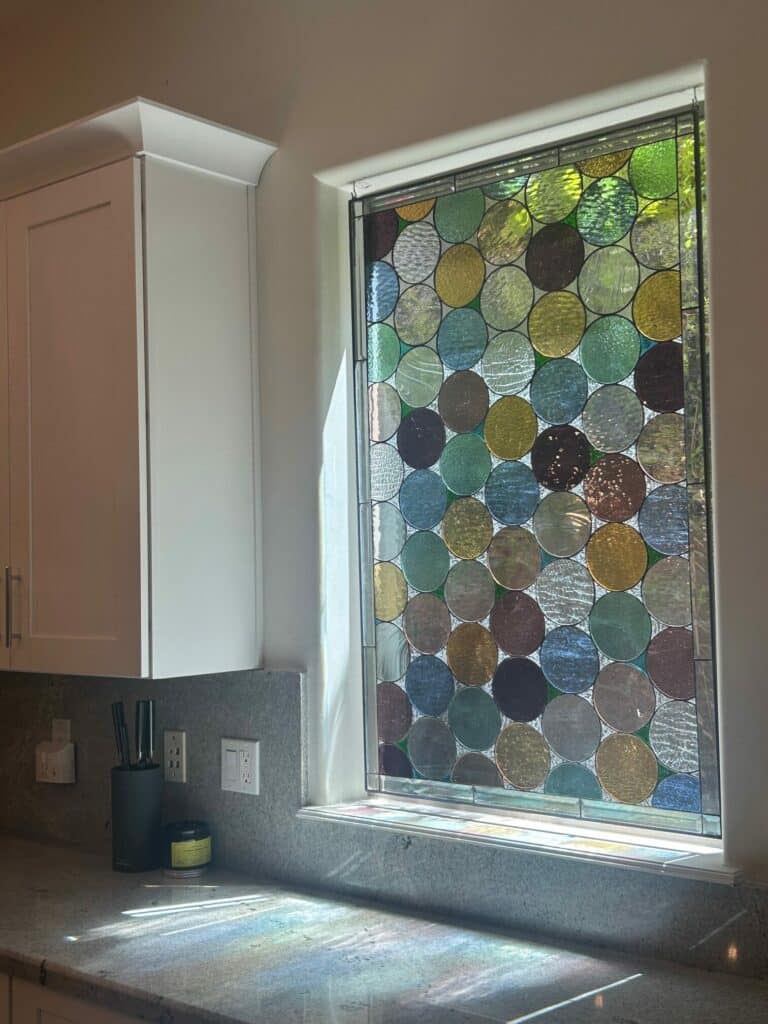 A rectangular leaded stained glass window panel featuring a dense mosaic of overlapping textured glass circles in vibrant shades of green, blue, yellow, amber, and brown, installed in a kitchen window above a speckled gray countertop.