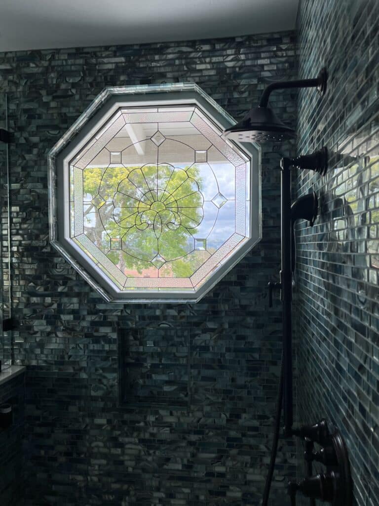 An octagonal leaded glass window installed in a modern shower with dark blue-green rectangular glass tiles. The clear, textured, and beveled glass forms a geometric pattern with a central sunburst or floral medallion. Outside the window, green tree foliage and a bright sky are visible, while dark bronze shower fixtures are on the right side.