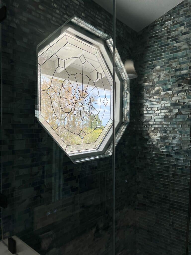 An octagonal, white-framed insulated window with a clear leaded glass panel, featuring a central floral medallion with beveled and textured clear glass. The window is installed in a modern shower with variegated blue and grey glass subway tiles, offering a view of green trees and blue sky outside.