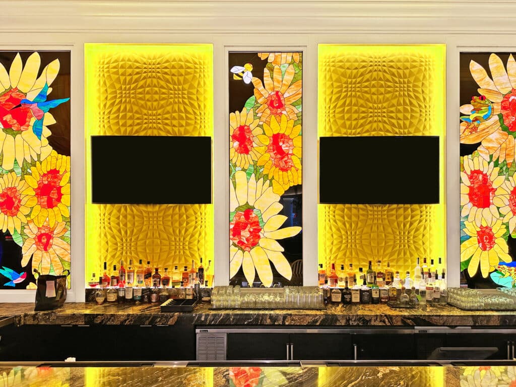 A vibrant bar installation featuring multiple tall stained glass panels depicting large sunflowers with red and yellow petals, green leaves, and various garden creatures like a hummingbird, bee, and frog. These panels alternate with bright yellow, textured, illuminated wall panels, each with a black rectangular screen mounted in the center. The foreground shows a dark marble bar top laden with liquor bottles and glassware.